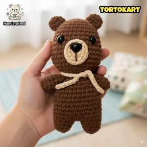 Hand holding a small handcrafted crochet teddy bear plush toy with black button eyes and beige accents, made by Tortokart